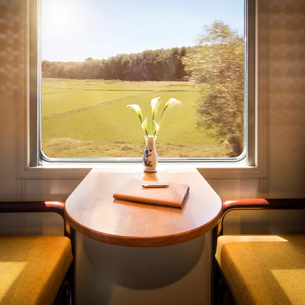 Sunlit window scene onboard the The Vietage by Anantara in Vietnam, with a small table and two seats. A vase with white flowers and a closed book sits on the table. Outside, lush green fields and trees blur past under a clear, blue sky.