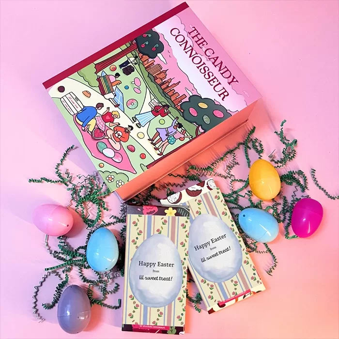 A colorful candy box labeled The Candy Connoisseur sits on a pink surface surrounded by pastel Easter eggs, green decorative grass, and two Happy Easter chocolate bar packages with egg-shaped windows.