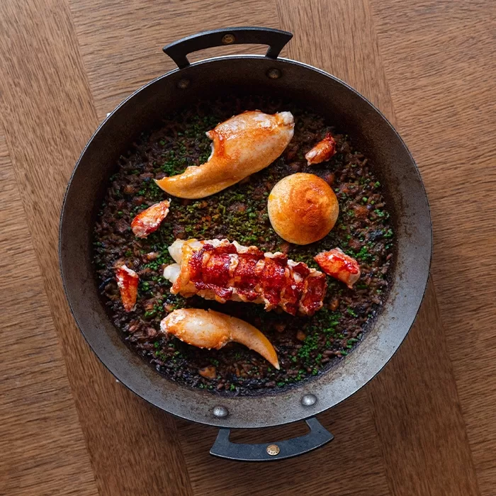 A black pan on a wooden table contains a gourmet dish with lobster tail, lobster claws, a round pastry, and finely chopped green herbs over a bed of dark grains or rice.