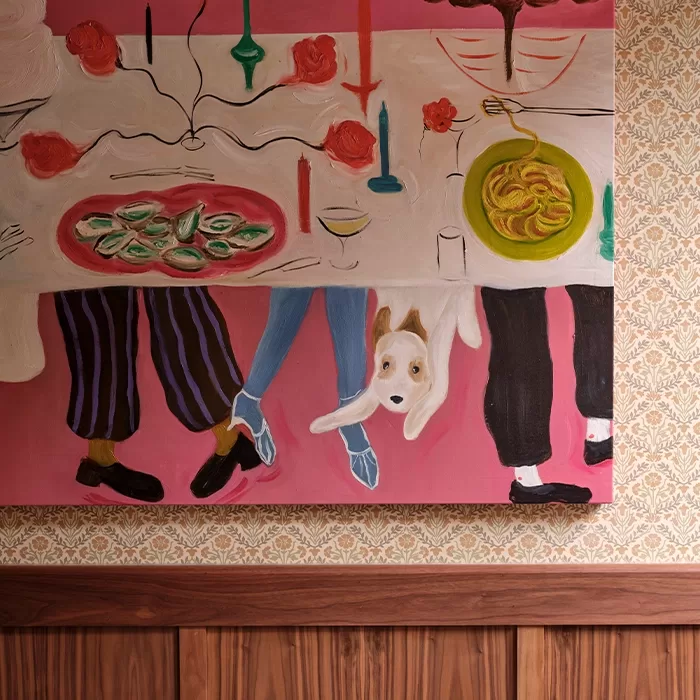 A colorful painting shows a dinner table with oysters, pasta, wine, and candles, viewed from above. Below the table, three people’s legs and a white dog peeking out are visible against a pink background.