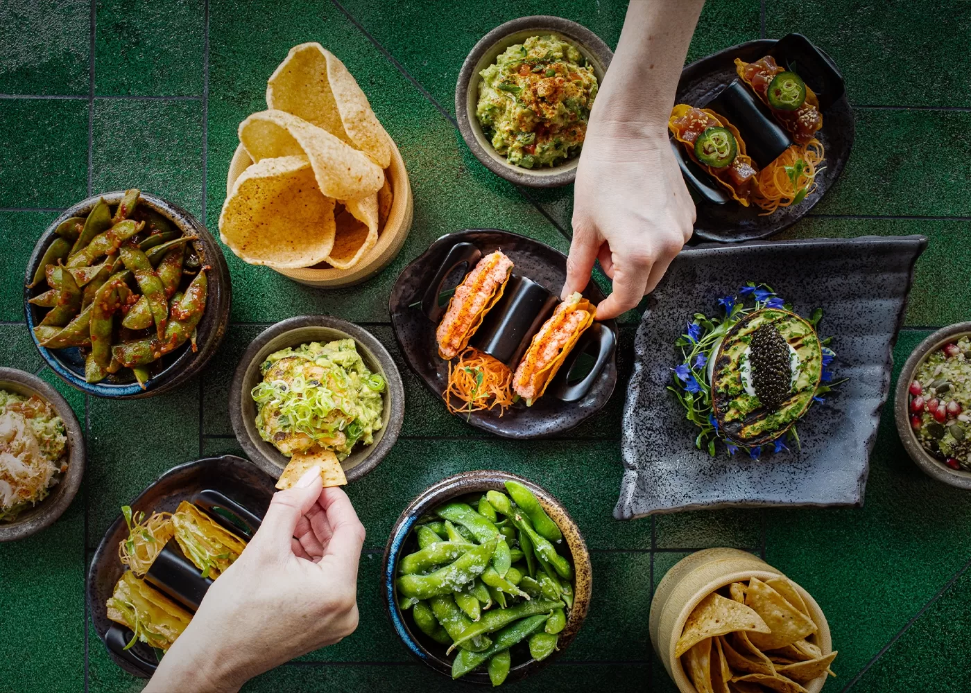 Several dishes of assorted Asian and Mexican-inspired appetizers, including sushi rolls, edamame, guacamole, and tortilla chips, are arranged on a green table as two hands reach in to serve themselves.