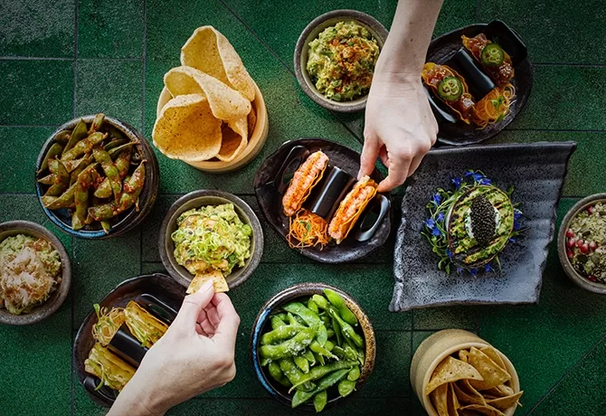 An overhead view of assorted dishes, including edamame, guacamole, tacos, seaweed rolls, chips, and various appetizers, with two people reaching in to grab food from the plates on a green-tiled surface.