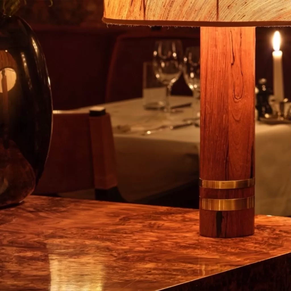 A wooden lamp with a beige shade sits on a polished brown table. In the background, a set dinner table with wine glasses and a lit candle create a warm, elegant atmosphere.