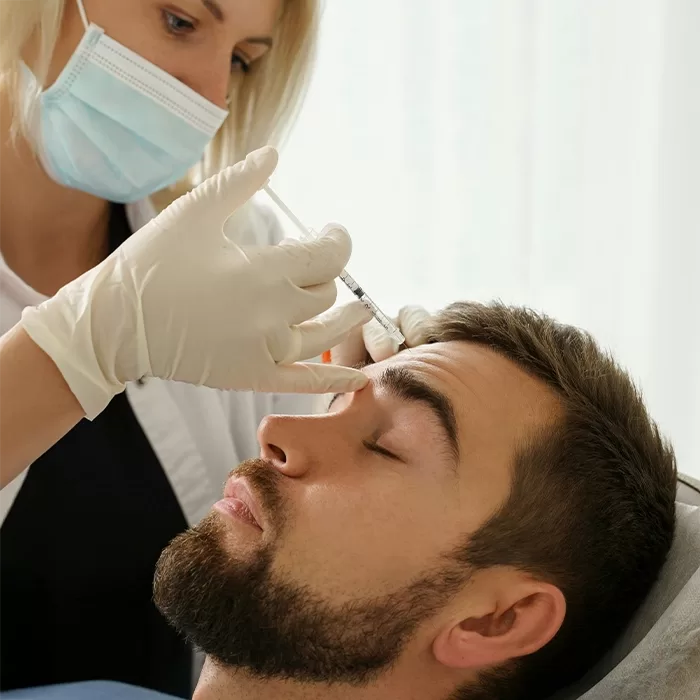 A person wearing gloves and a face mask administers an injection to a mans forehead as he lies down with his eyes closed, possibly during a cosmetic or medical procedure.