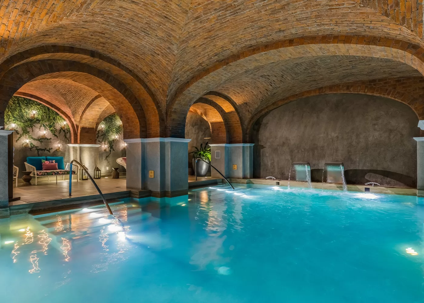 An elegant indoor swimming pool under arched brick ceilings, with water features, steps leading into the pool, lounge chairs, greenery, and soft ambient lighting creating a relaxing atmosphere.