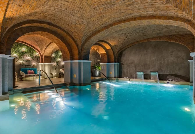 A luxurious indoor pool with arched brick ceilings, soft lighting, and cascading water features. Relaxation areas with sofas and plants are set along the sides, creating a tranquil spa atmosphere.