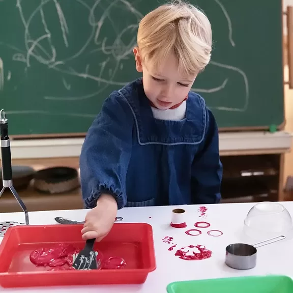 A young child in a blue smock uses a spatula to spread red paint on a tray at a table. Various art supplies are on the table, and a chalkboard with scribbles is in the background.