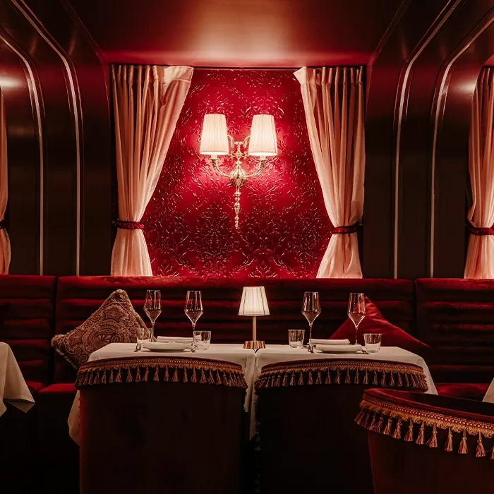 Lavish restaurant interior with red velvet seating, ornate pillows, and tassel-trimmed chairs. Tables are set with wine glasses and a small lamp. Red wallpaper and draped curtains frame a decorative wall sconce with two lights.