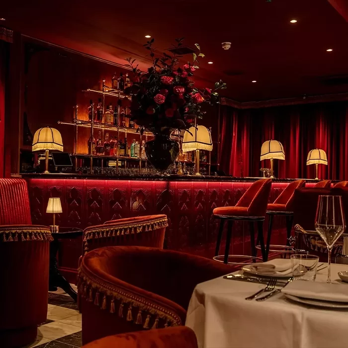 Elegant, dimly lit restaurant and bar with red velvet chairs, a marble-topped table set for dining, lamps with warm light, a large flower arrangement, and shelves of liquor bottles behind the bar.
