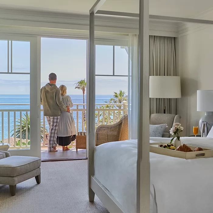 A couple stands on a balcony overlooking the ocean from a bright, elegant hotel room with a four-poster bed, breakfast tray, and modern decor.