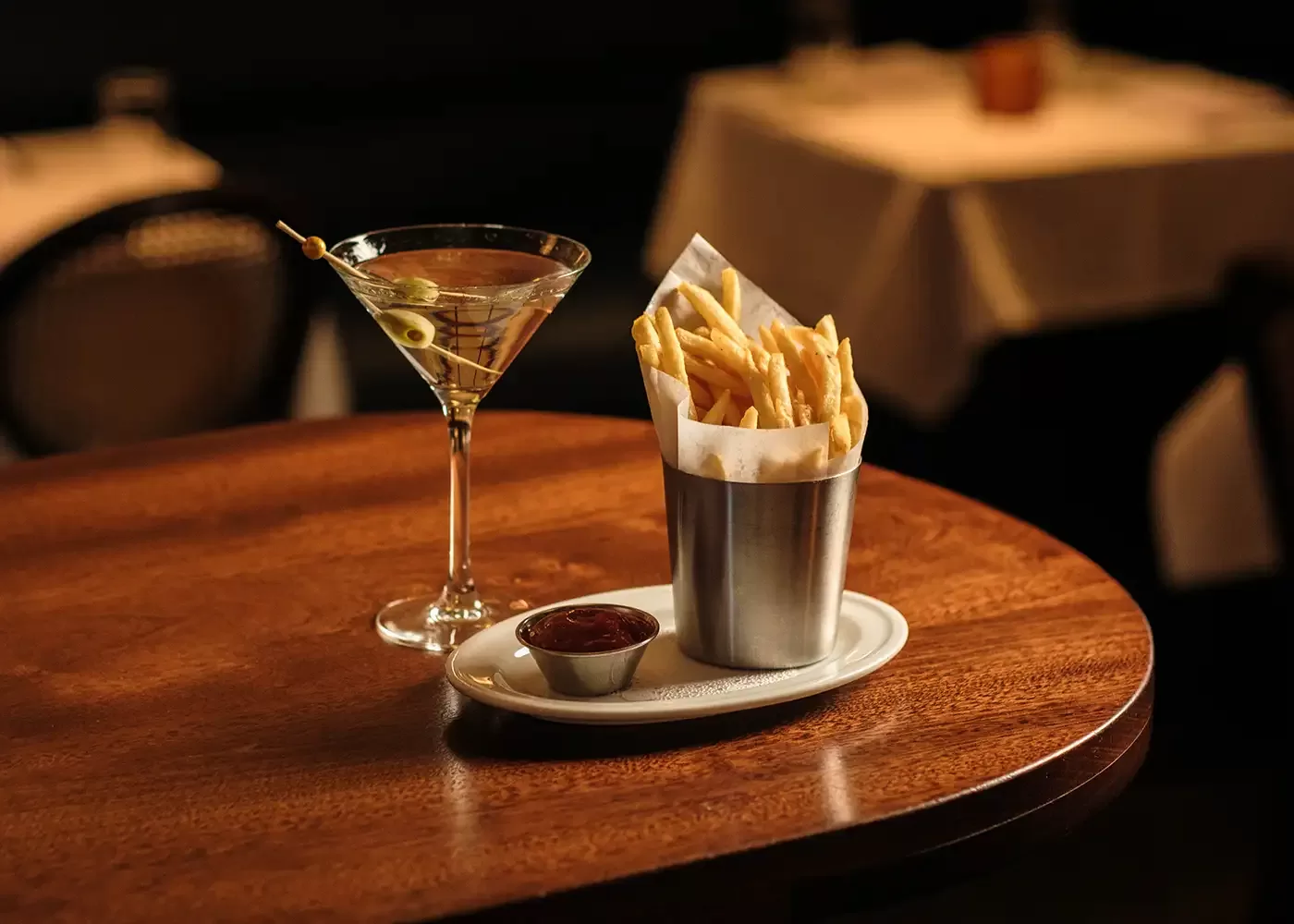 A glass of martini with olives and a metal cup filled with French fries, served with ketchup on a small white plate, sitting on a wooden table in a dimly lit restaurant.