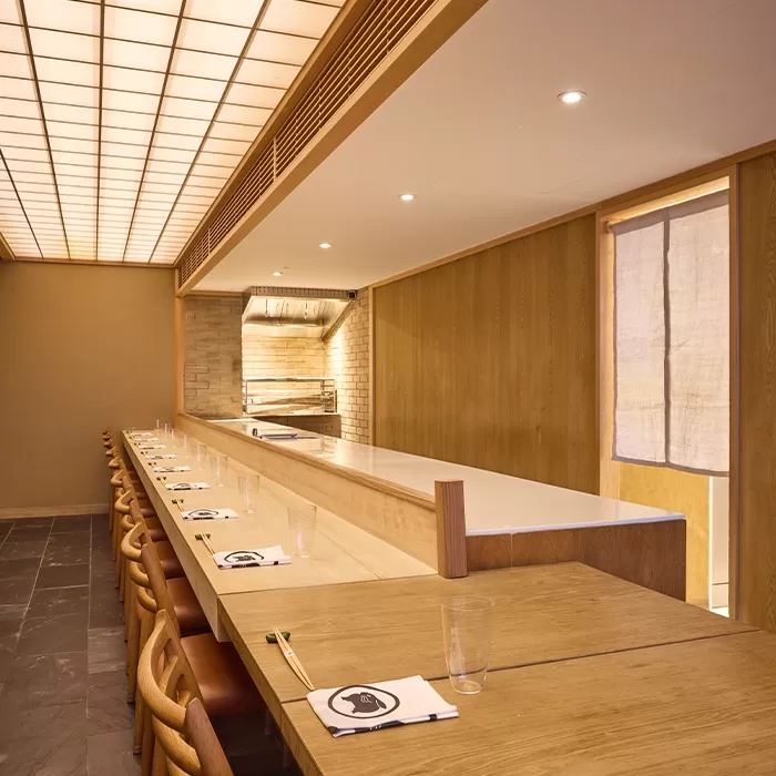 Minimalist Japanese restaurant interior with a long wooden counter, simple light wood chairs, neatly arranged napkins, chopsticks, and glasses. Soft lighting and clean lines create a calm, inviting atmosphere.