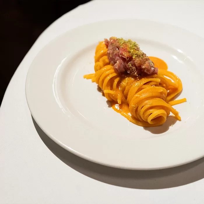A neatly twirled serving of orange-colored pasta topped with a finely chopped pink mixture sits on a white plate, placed on a white tablecloth.
