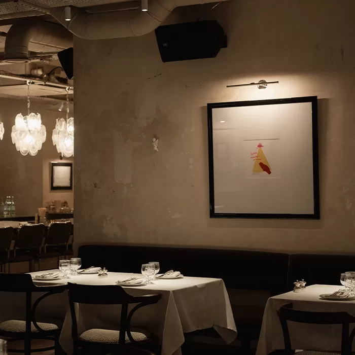 A dimly lit, elegant restaurant with white tablecloths, neatly set tables, and dark wooden chairs. Modern light fixtures and framed abstract art decorate the beige walls, creating a cozy, intimate atmosphere.
