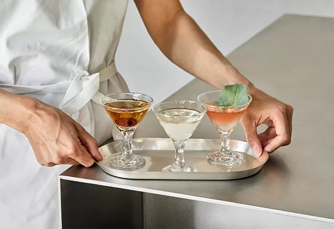 A person in a white apron holds a silver tray with three different cocktails in small martini glasses, each with a distinct color and garnish, placed on a gray countertop.