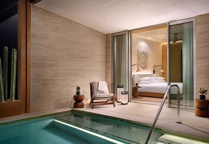 A modern hotel room with sliding glass doors opens to a private patio featuring a small pool, a lounge chair with a cushion, side tables with potted plants, and soft ambient lighting.
