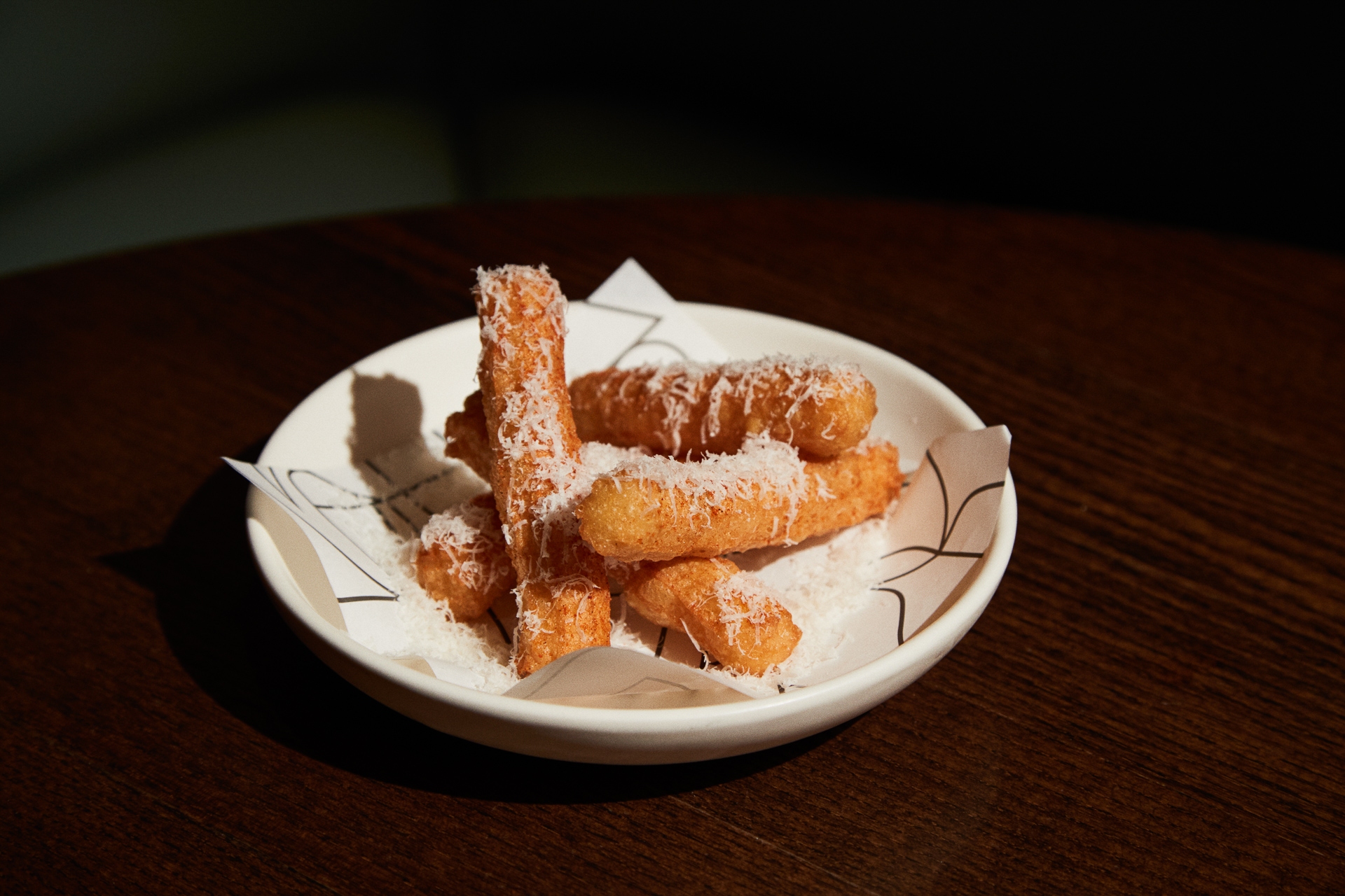 A plate of churros dusted with grated cheese, served on a wooden table. The churros sit on a white plate lined with paper, with a moody lighting casting shadows at Luca, Farringdon.