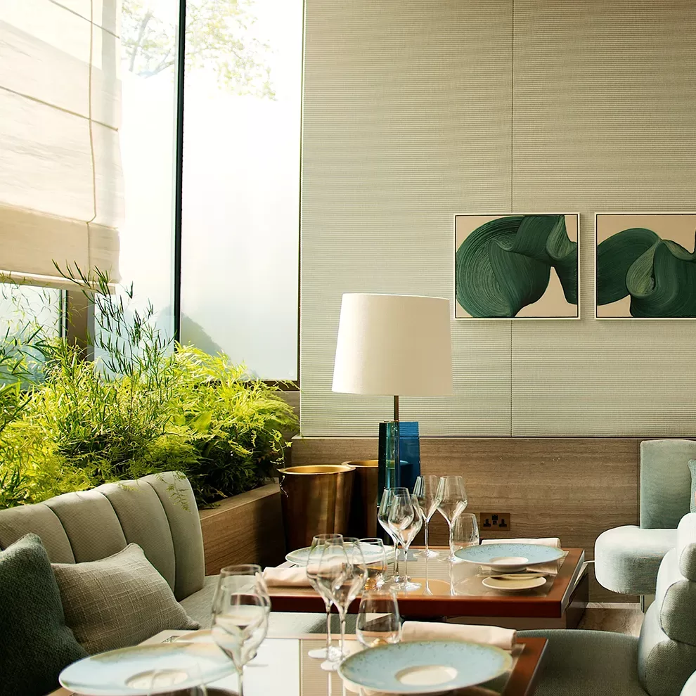 A modern, elegantly decorated dining room with a set table. The room features minimalist artwork, a table with glasses and plates, a lamp, and a large window with greenery outside. Neutral tones and natural light create a serene atmosphere.