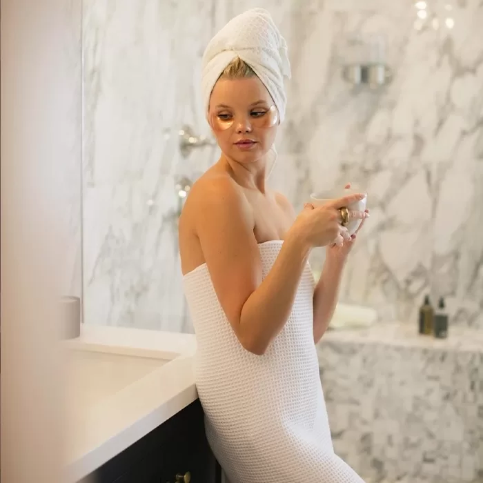Woman with a towel wrapped around her body and hair sits in a marble bathroom, wearing under-eye patches and holding a cup, looking to the side.