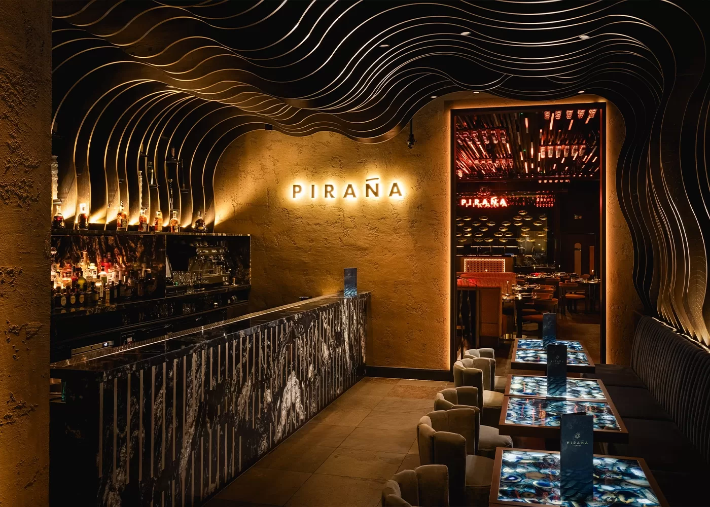 A stylish, dimly lit bar with wavy sculpted ceiling, illuminated “PIRAÑA” sign on a textured wall, a sleek bar counter, and tables with blue lighting, creating a modern, upscale atmosphere.