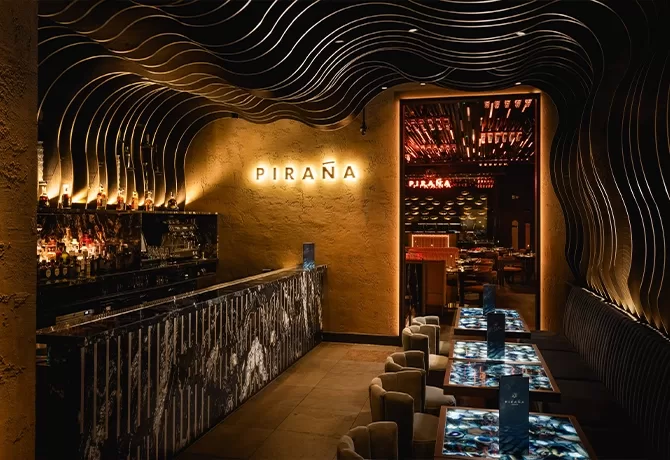 Modern restaurant interior with a wavy, sculptural ceiling, a long bar on the left, and dim lighting. The word PIRAÑA is illuminated on the back wall. Elegant tables and chairs are set for dining.