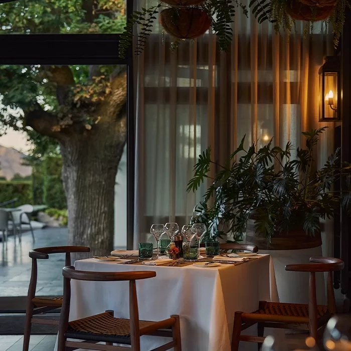 A warmly lit restaurant dining table set for four, with wooden chairs and a white tablecloth, sits near large windows overlooking a patio and lush greenery outside.