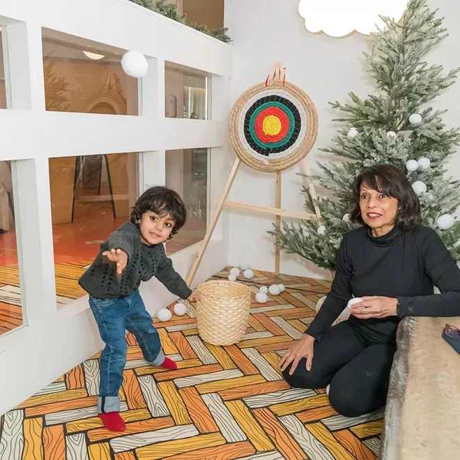 A child in a gray sweater throws a snowball at a target while a woman in a black outfit sits beside him, holding a snowball. They are indoors near a Christmas tree, with snowballs scattered around and a colorful target on an easel.