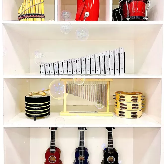 A neatly organized white shelf displaying colorful musical instruments, including xylophones, bongos, tambourines, ukuleles, and a chime. Soap bubbles float in front of the shelves, adding a playful touch.