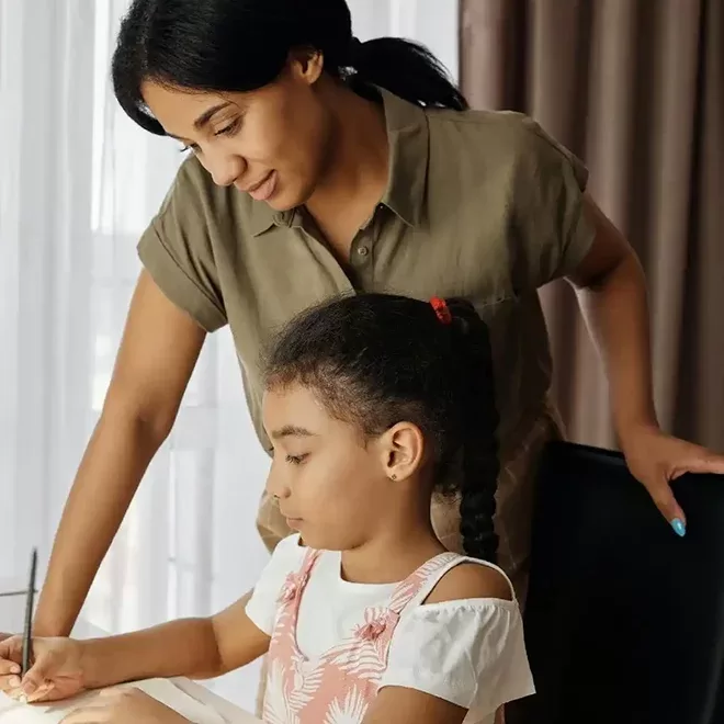 A young girl is seated at a table, focused on writing on a sheet of paper. She wears a white T-shirt under a light pink dress with floral patterns. A woman, likely her mother or teacher, stands beside her, smiling warmly as she offers guidance. The setting is cozy, with natural light filtering through the curtains in the background.