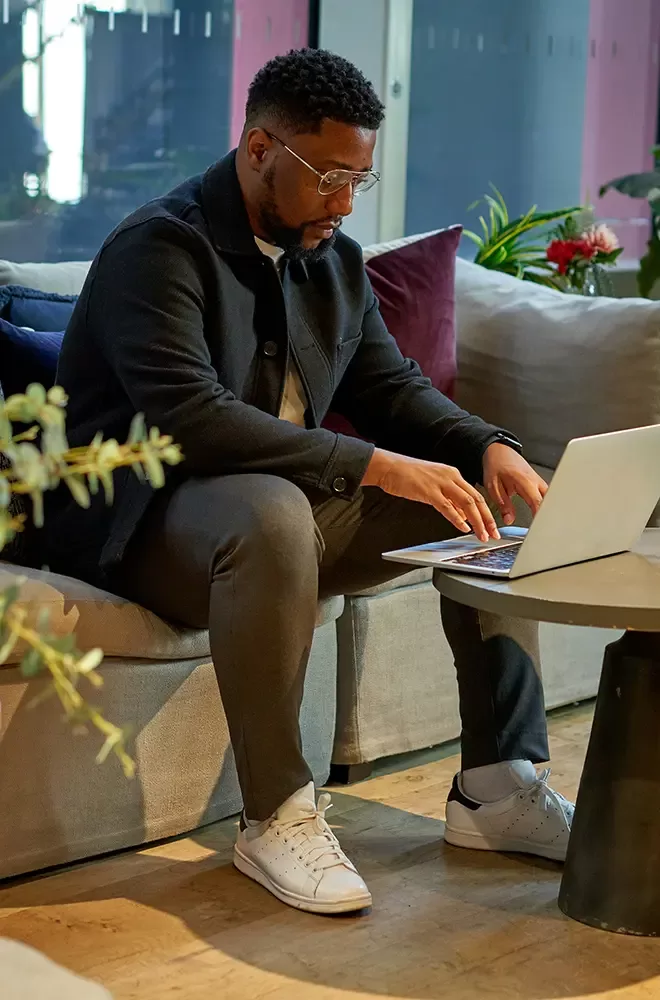 A man sitting on a comfortable sofa in a modern, cozy room, working on a laptop placed on a round coffee table. The room is stylishly decorated with plush cushions, indoor plants, and a large window in the background. The man is focused on his work, wearing casual attire, including a black jacket, dark pants, and white sneakers.