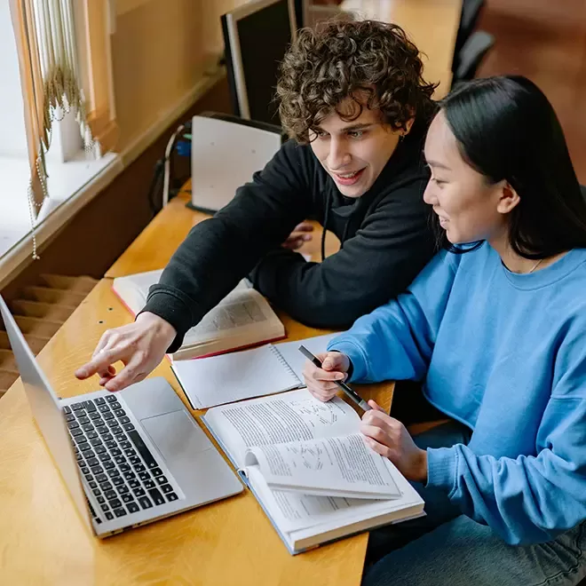 Two students sit at a wooden desk in a library or study area. They are looking at a laptop screen, with books open in front of them. The male student with curly hair points at the screen while the female student, wearing a blue sweater, holds a pen and smiles. Both are engaged in a collaborative study session.