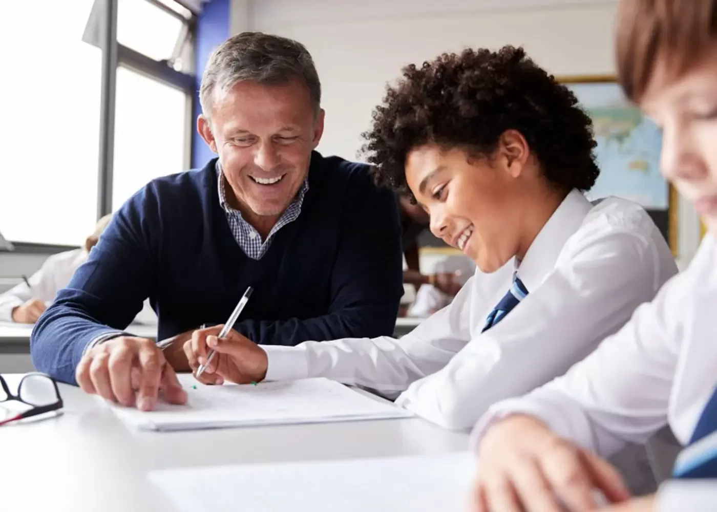 Male teacher and students smiling, sitting at desk in a school completing work with a pen and paper.