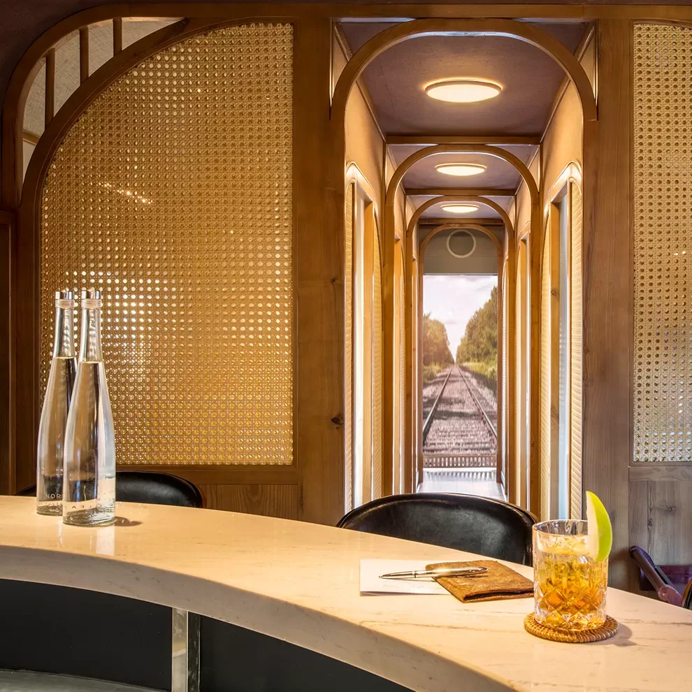 Cosy interior of The Vietage by Anantara in Vietnam, with a curved bar countertop holding two water bottles, a notebook, and a drink garnished with a lemon slice. The background features wooden partitions and a narrow corridor leading to an artistic backdrop of train tracks.