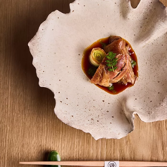 A gourmet dish featuring sliced meat, a green vegetable, and sauce, artfully arranged on a textured, cream-colored ceramic plate. Wooden chopsticks rest on the table beside the plate.
