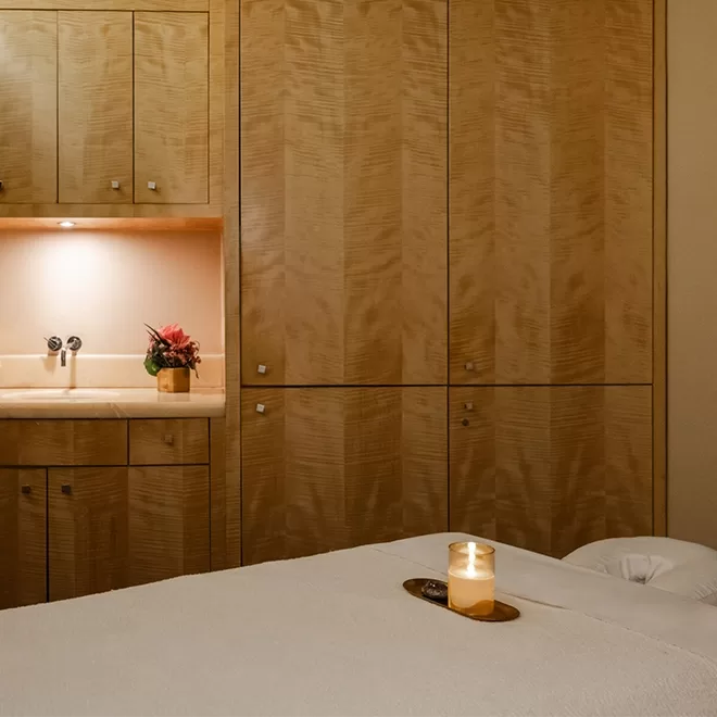 A tranquil spa room with a massage table covered in a white sheet, a lit candle on a wooden tray, wooden cabinets, a built-in sink, and a small flower arrangement under warm lighting.
