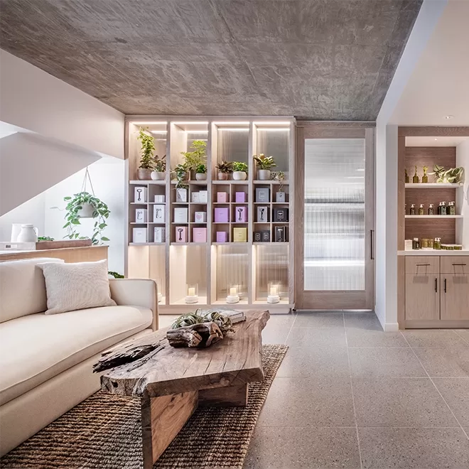 Modern living space with a cream sofa, rustic wooden coffee table, and built-in shelves displaying boxes, jars, and potted plants; warm lighting and neutral tones create a cozy, inviting atmosphere.