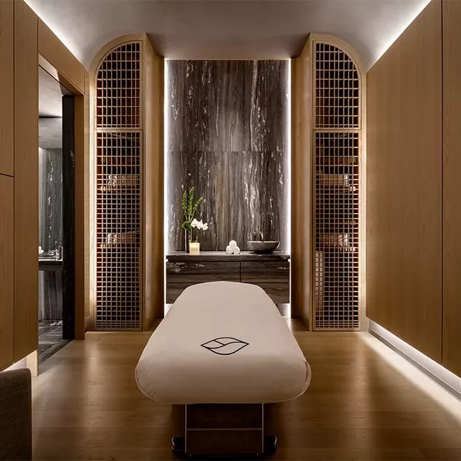 Luxurious spa room with a massage table in the center, elegant wooden walls, soft lighting, and decorative shelving. A vase with white flowers and a bowl sit on a marble counter at the back.