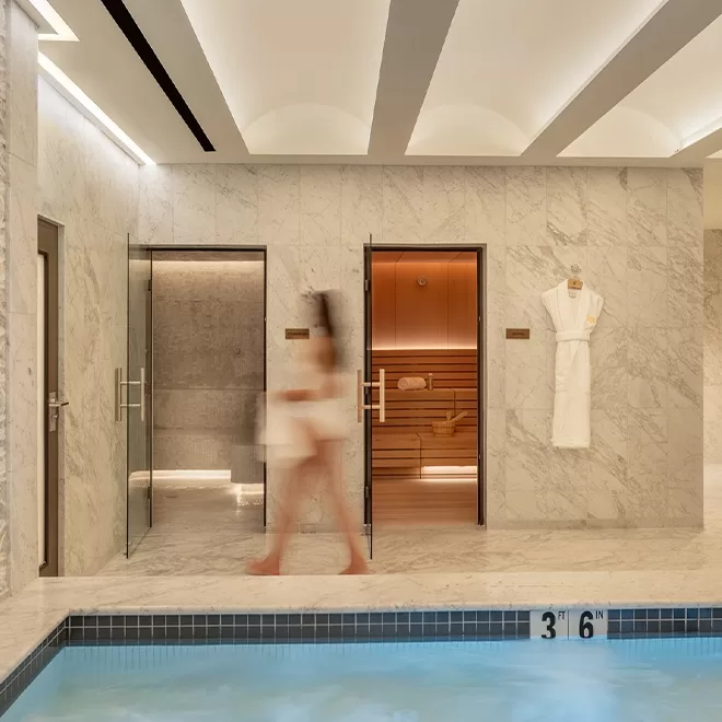 A blurred person in a towel walks past a marble-walled spa area with glass doors leading to a steam room and sauna; a white robe hangs on the wall beside the pool marked 3 6.