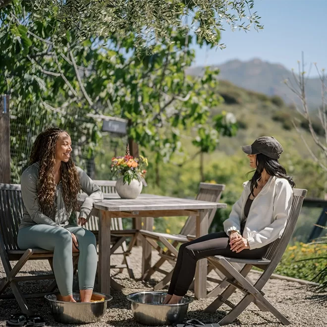 Two women sit on wooden chairs outdoors by a table, smiling and talking. Both have their feet in metal tubs of water. Trees and mountains are visible in the background on a sunny day.