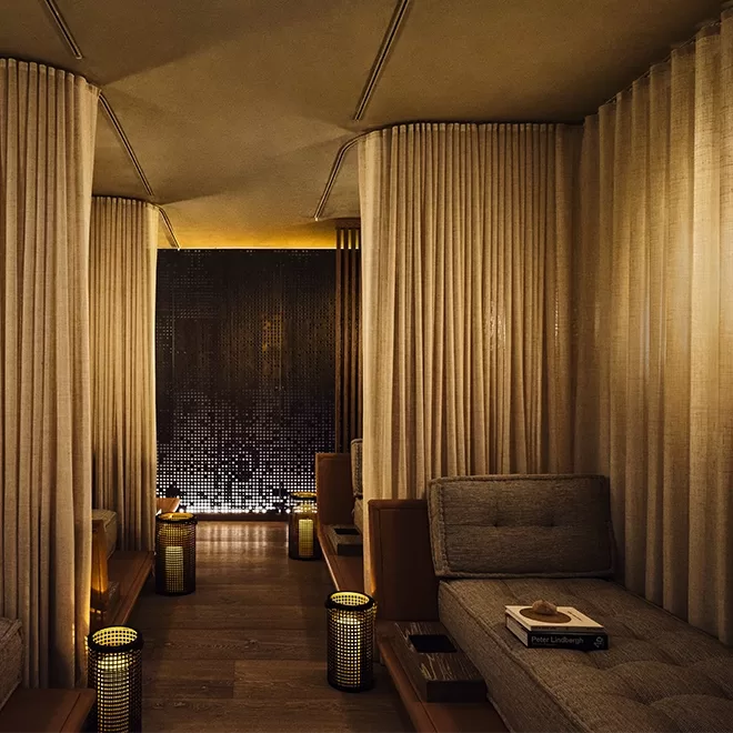 A cozy lounge area with soft lighting, beige curtains dividing private seating spaces, cushioned benches, wooden floors, and small lanterns creating a warm, inviting atmosphere.