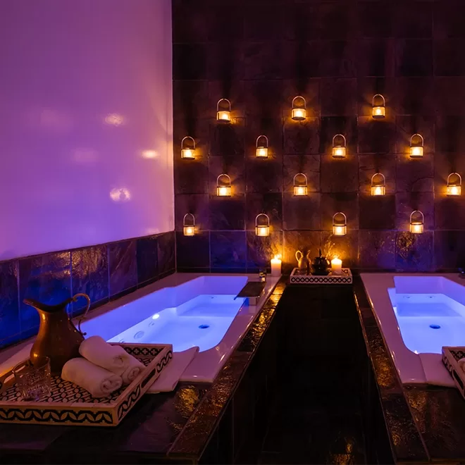 A dimly lit spa room with two glowing bathtubs, candles on the tiled wall, towels, and a decorative pitcher on a tray, creating a relaxing and luxurious atmosphere.