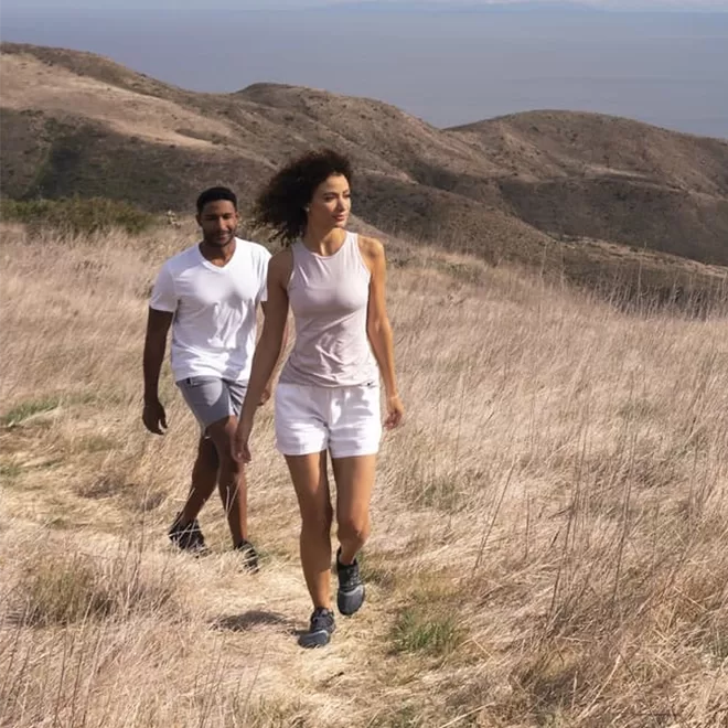 A woman and a man are hiking on a narrow dirt trail through dry grass and rolling hills, with the ocean visible in the distance under a clear sky.