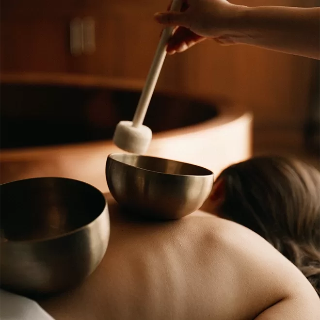 A person lies face down with two brass singing bowls placed on their back, while another hand holds a mallet above one of the bowls, preparing for a sound therapy session in a calming, softly lit room.