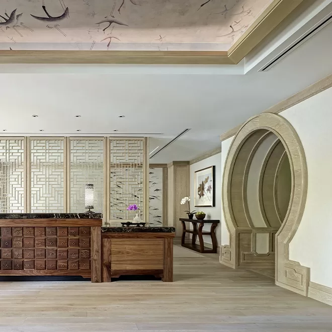 A modern room with Asian-inspired decor, featuring a wooden desk, lattice panel walls, a round moon gate doorway, and a ceiling with bird designs. Light wood floors and elegant furniture complete the serene space.