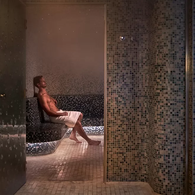 A man wrapped in a towel sits and relaxes on a tiled bench inside a steamy, mosaic-tiled steam room with a door open to the left.