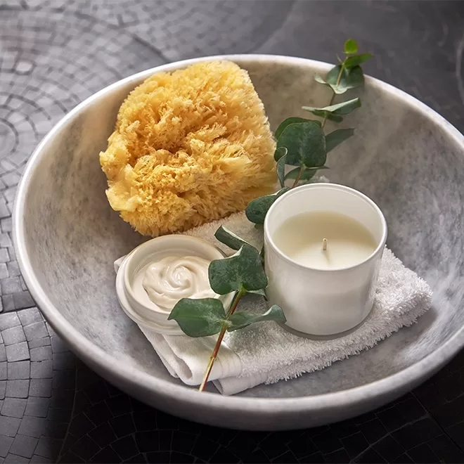 A marble bowl containing a natural sponge, a white candle in a glass holder, a small dish of cream, a white folded towel, and a green eucalyptus branch, set on a dark tiled surface.