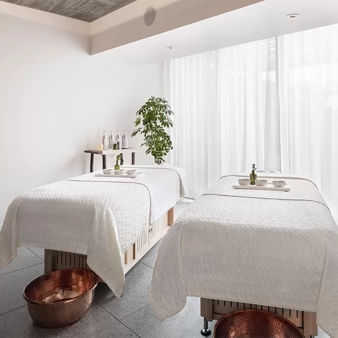 Two massage tables with white linens are set up side by side in a bright, modern spa room. Copper bowls are placed below each table, and a small table with bottles and a plant decorate the serene space.