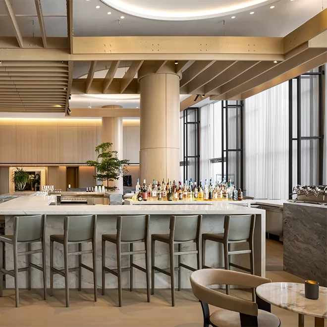 A modern, upscale bar with a marble counter, high-backed chairs, and a variety of liquor bottles. The space features tall windows, neutral tones, wood accents, and contemporary lighting.