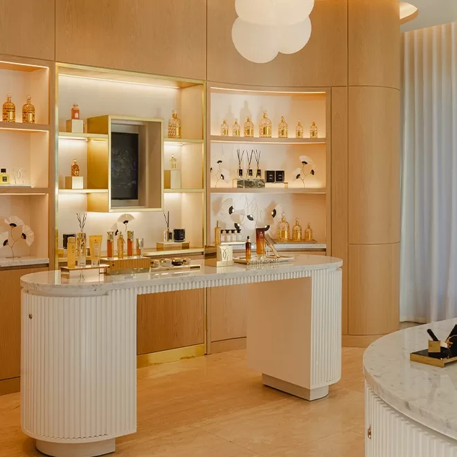 A modern, elegant perfume store with light wood shelves displaying various perfume bottles. A curved white marble table holds more fragrances, and soft lighting creates a warm, inviting atmosphere.