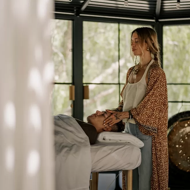 A woman in a patterned robe gently places her hands on a persons head, giving a massage or energy treatment in a bright, peaceful room with large windows.